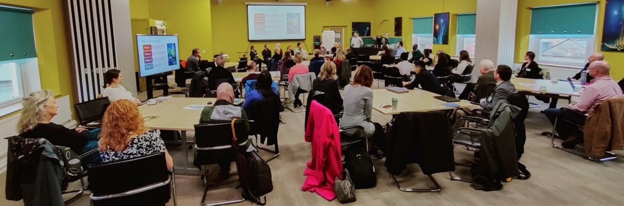 About 40 people listening to a presentation in a room with yellow walls
